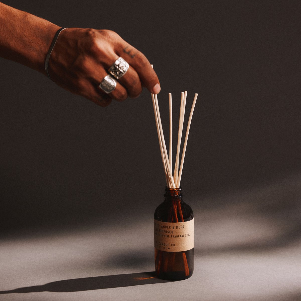 Hand with a ring holding reed diffuser sticks next to a bottle of amber & moss reed diffuser on a dark background