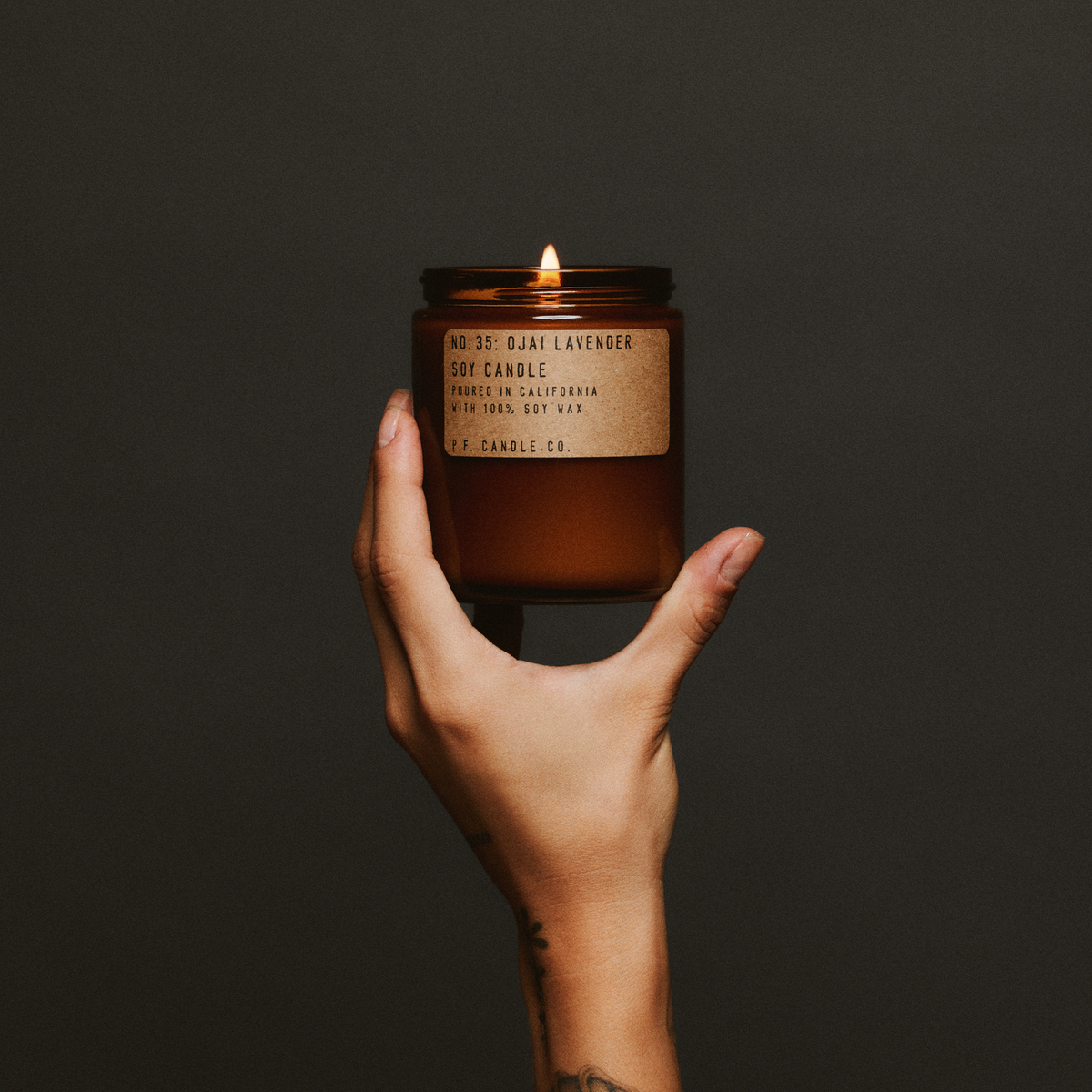 Hand holding a lit ojai lavender standard candle with a label against a dark background