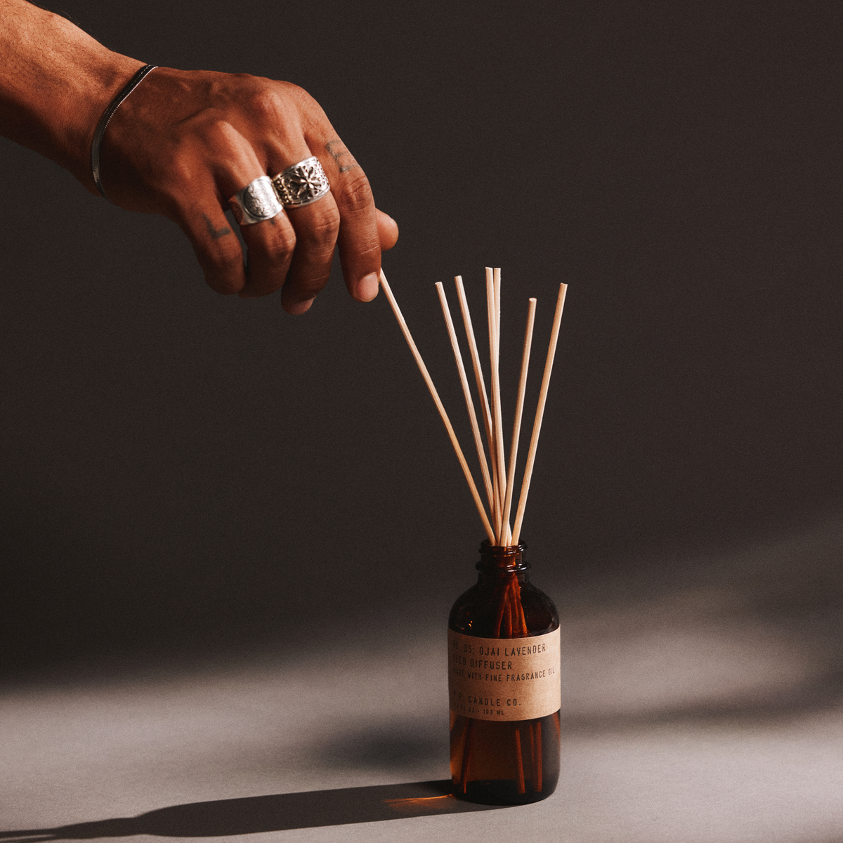 Hand with a ring holding reed diffuser sticks next to a bottle of ojai lavender reed diffuser on a dark background