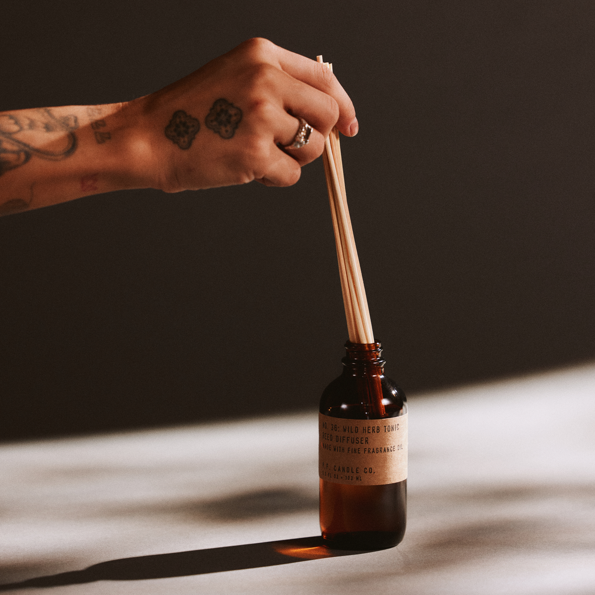 Hand with a ring holding reed diffuser sticks next to a bottle of wild herb tonic reed diffuser on a dark background